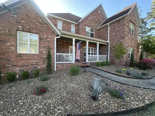 A river-rock landscape bed with a boulder, new plantings and a curved walk in front of a two-story brick house