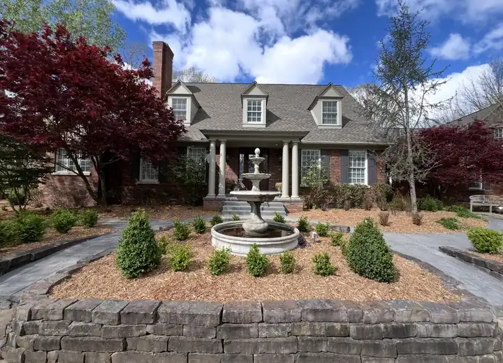 A circular front bed with a tiered stone fountain, fresh mulch, clipped boxwoods and a stacked-stone retaining wall in front of a brick colonial home